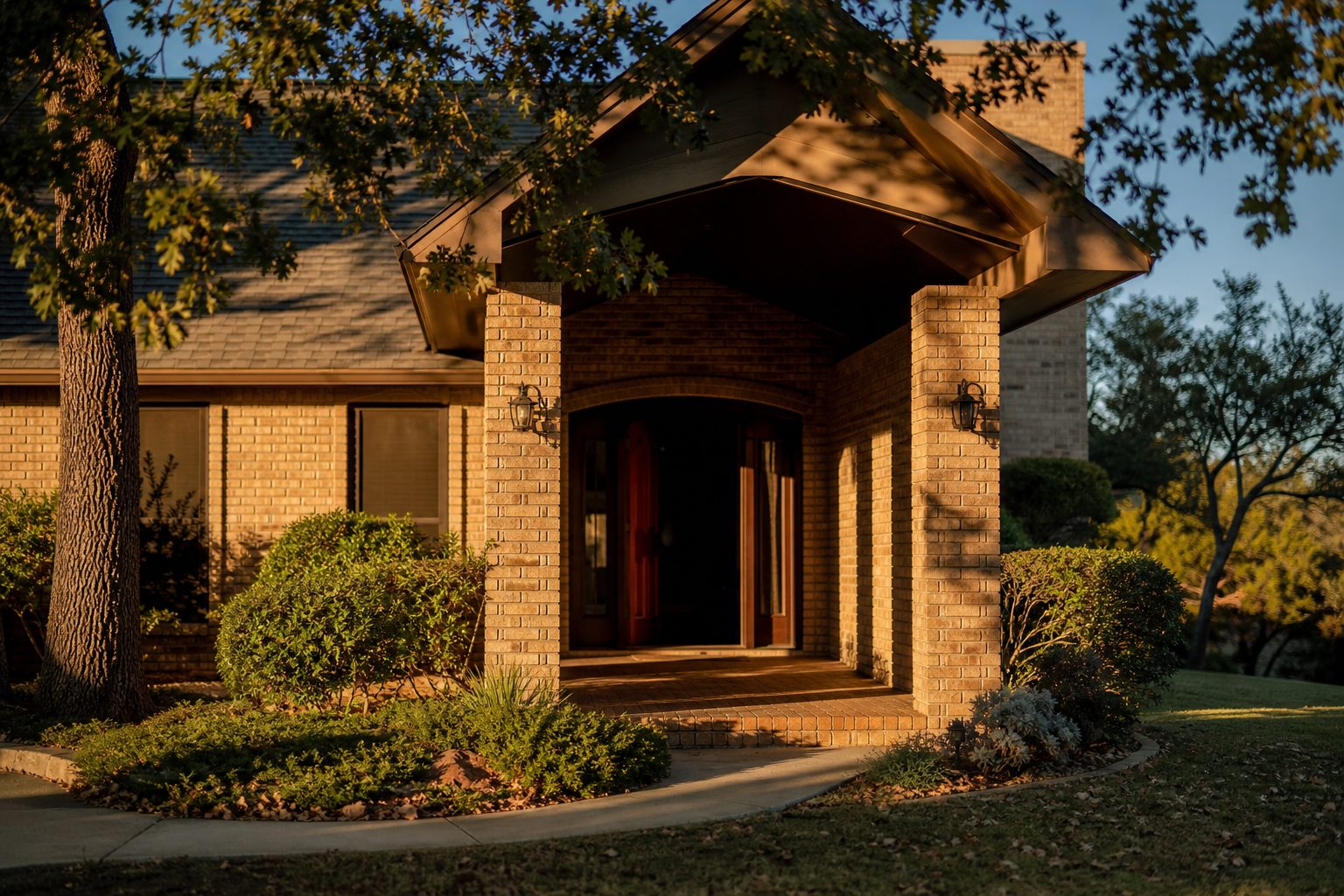 Front entrance and portico of brick home in Springtown with mature oak trees and golden afternoon light.
